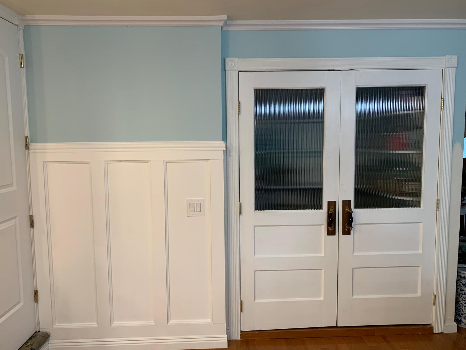 After photograph showing white wall paneling and repurposed antique pantry doors with ribbed glass and brass hardware
